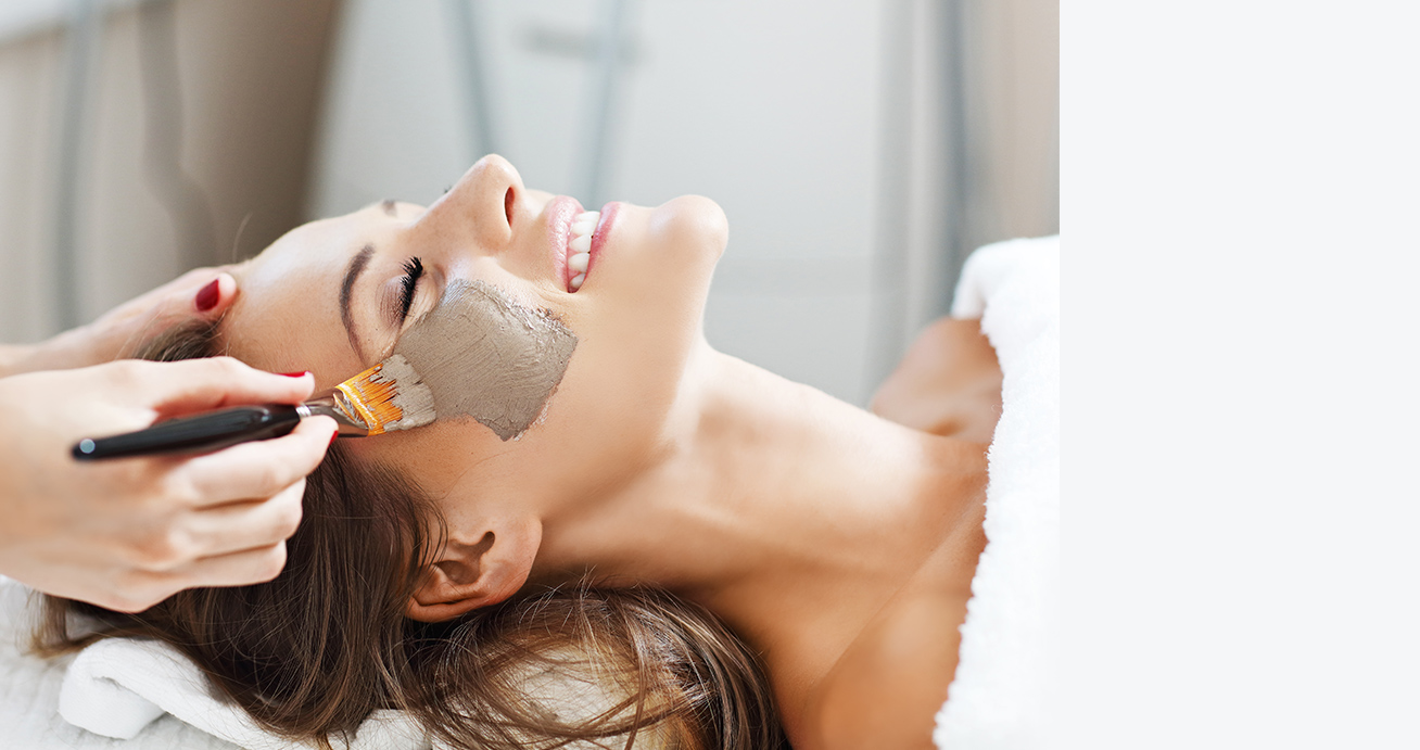 A smiling woman lies face-up as a facial skincare treatment is applied to her face with a brush